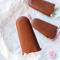 Hand holding a chocolate-covered cottage cheese popsicle on a unicorn stick over parchment paper