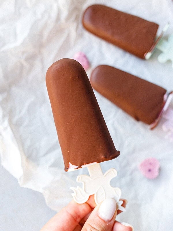 Hand holding a chocolate-covered cottage cheese popsicle on a unicorn stick over parchment paper