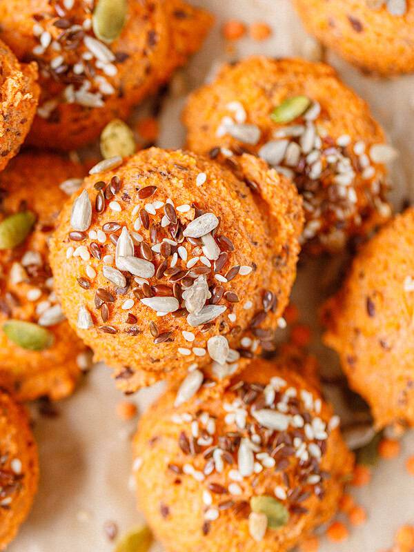 Red lentil bread rolls topped with sesame, flax, and pumpkin seeds, stacked on parchment paper