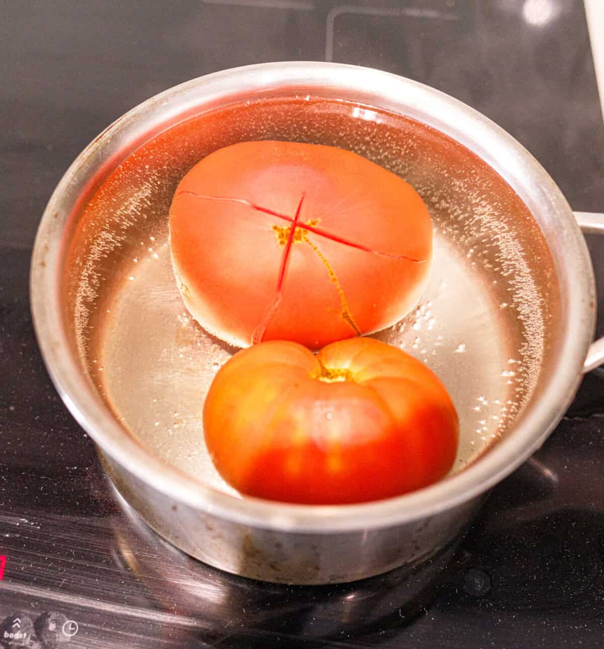 Two scored tomatoes blanching in a pot of boiling water on the stove