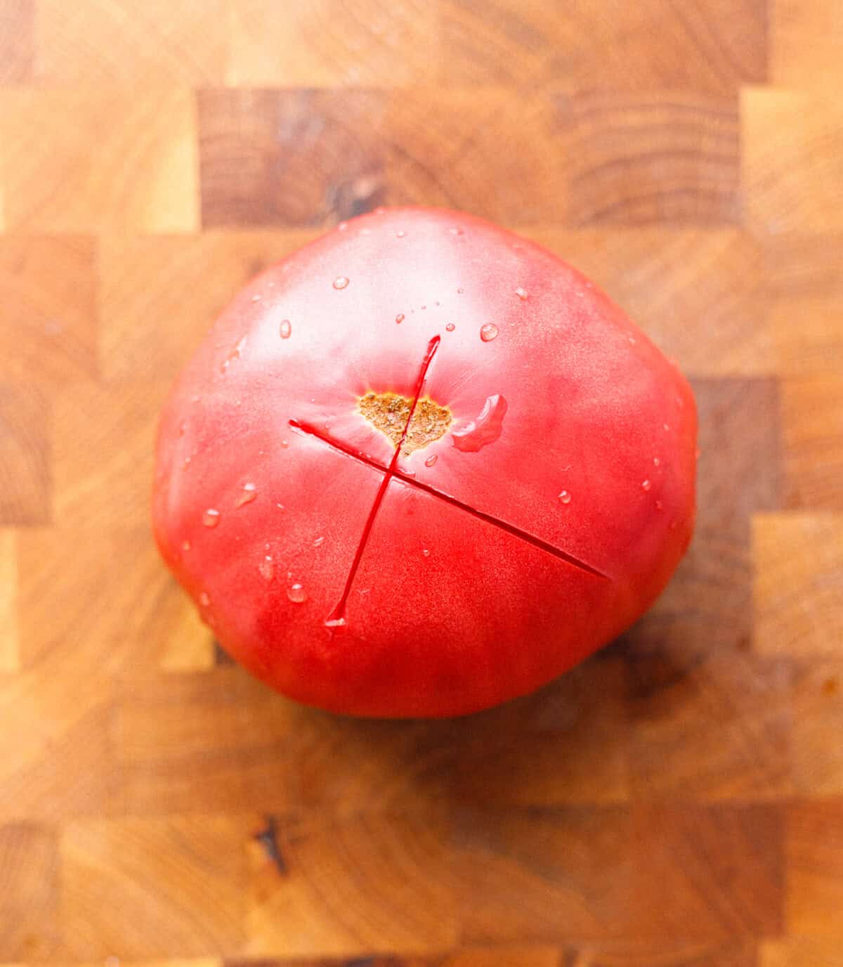 Tomato scored with an X on the bottom on a wooden cutting board before blanching