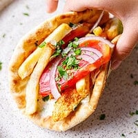 Close-up of a chicken gyros pita wrap held in one hand, loaded with chicken, fries, tomato, red onion and fresh parsley