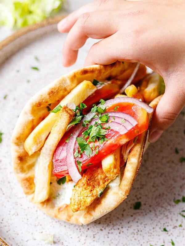 Close-up of a chicken gyros pita wrap held in one hand, loaded with chicken, fries, tomato, red onion and fresh parsley