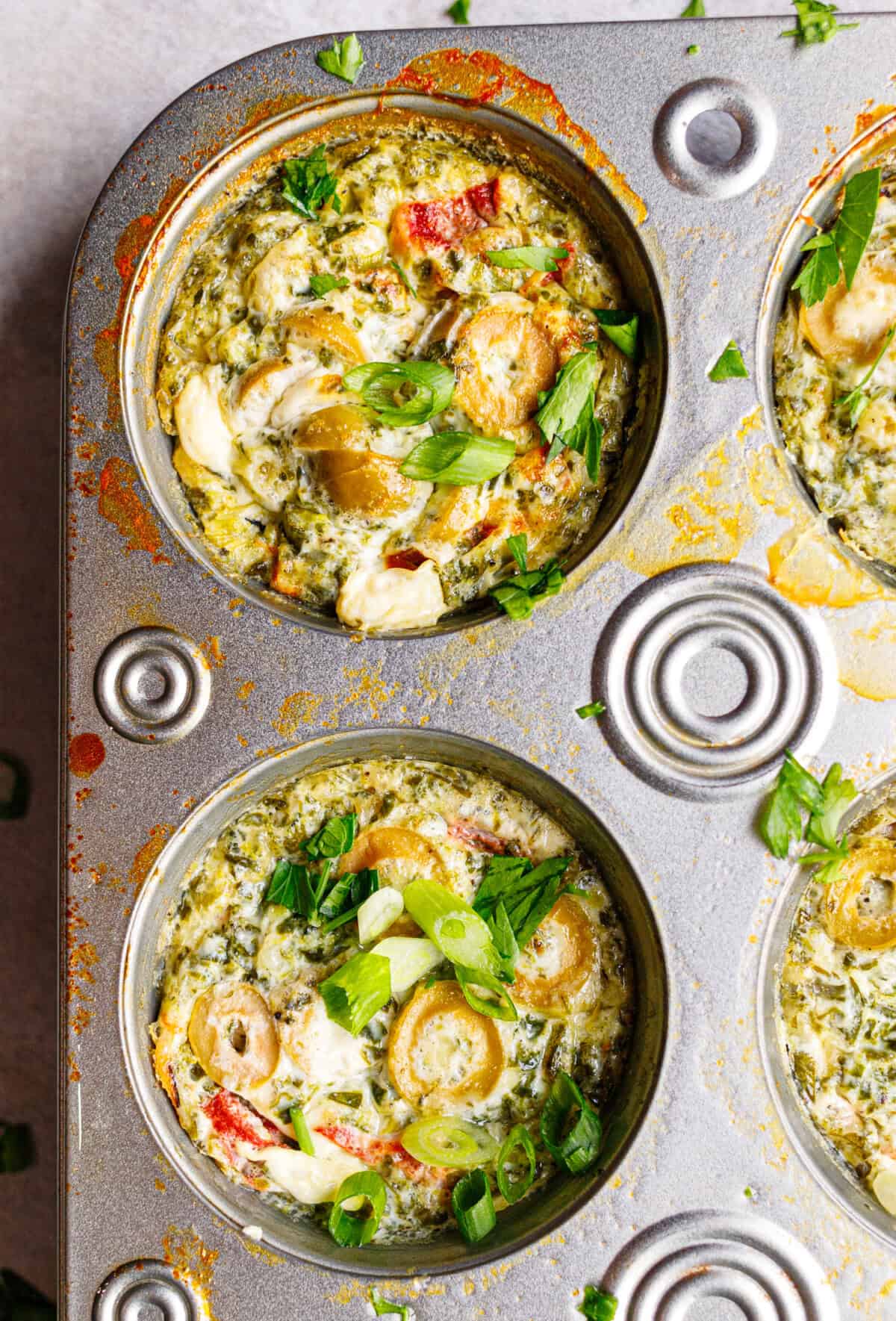 Two baked egg white cups in a muffin tin topped with green onion and parsley, edges golden and set