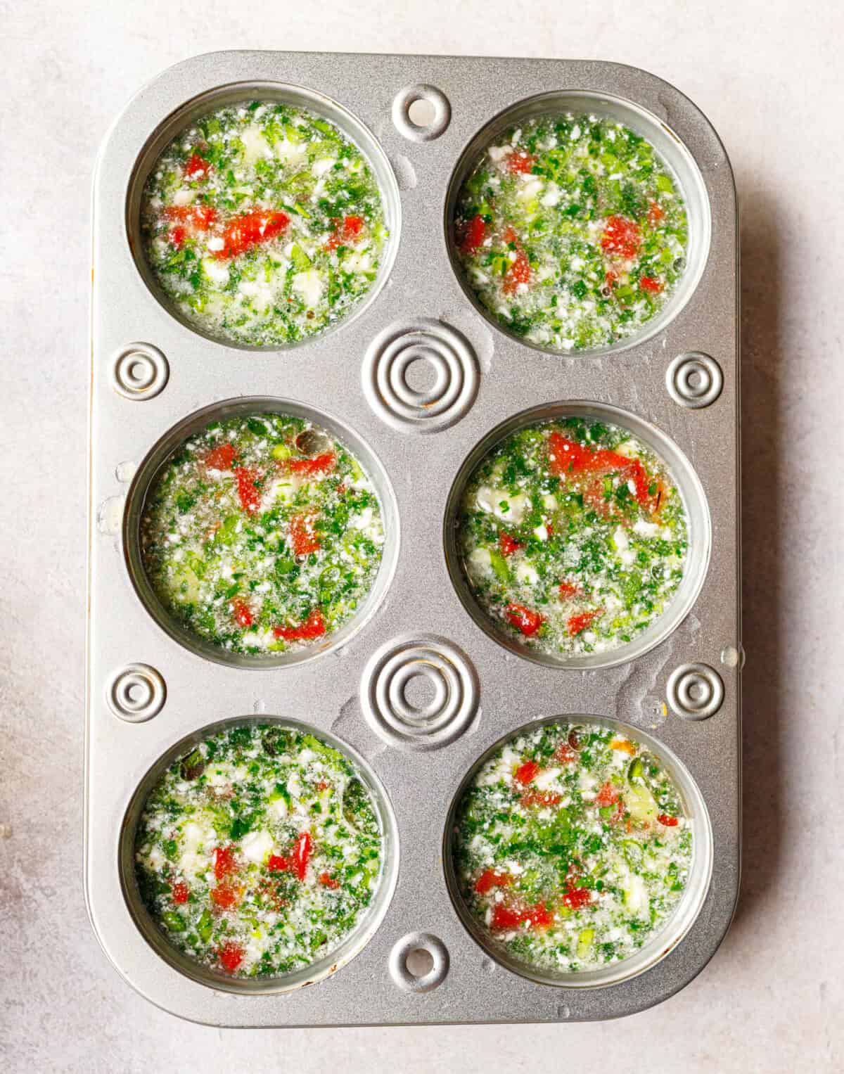 Muffin tin filled with raw egg white mixture, spinach, roasted red pepper and feta before baking