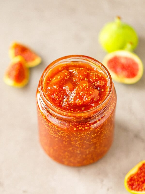 Glass jar of chunky fig jam full of tiny seeds, with halved green figs in the background