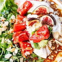 Gold fork lifting tomato, fig and arugula from an arugula burrata salad, with balsamic glaze pooled in the torn burrata