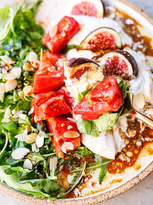 Gold fork lifting tomato, fig and arugula from an arugula burrata salad, with balsamic glaze pooled in the torn burrata