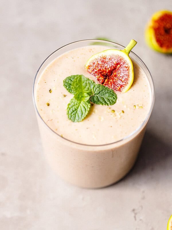 Glass of pale pink fig smoothie with a green fig wedge and mint on a gray concrete surface