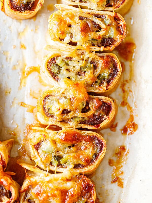Baked ground beef tortilla pinwheels on parchment paper, cut side up, with melted browned cheese on top and a spiral of beef, chopped pickles and cheese inside.