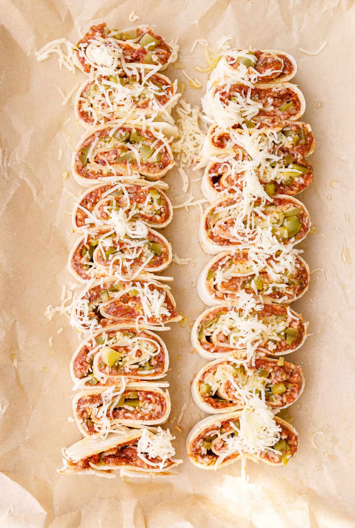 Two rows of unbaked ground beef tortilla pinwheels on parchment paper, cut side up, topped with grated white cheese.