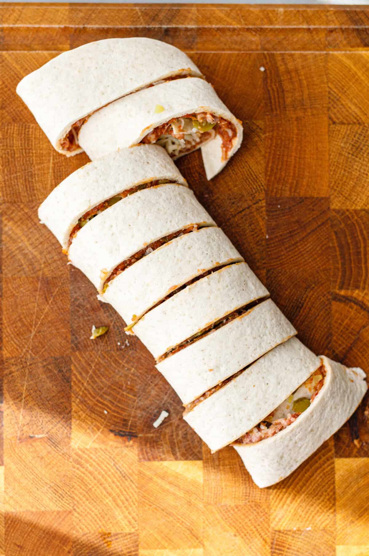 A rolled tortilla filled with ground beef, pickles and cheese, sliced into thick rounds on a wooden cutting board.