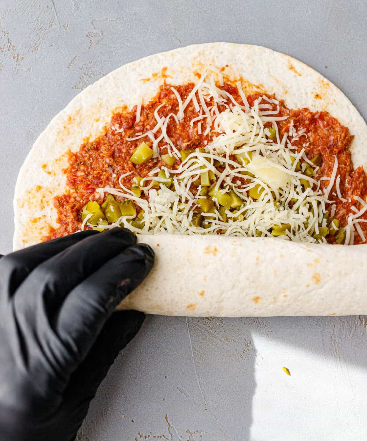 A gloved hand rolling up a flour tortilla spread with raw seasoned ground beef, chopped pickles and grated cheese.