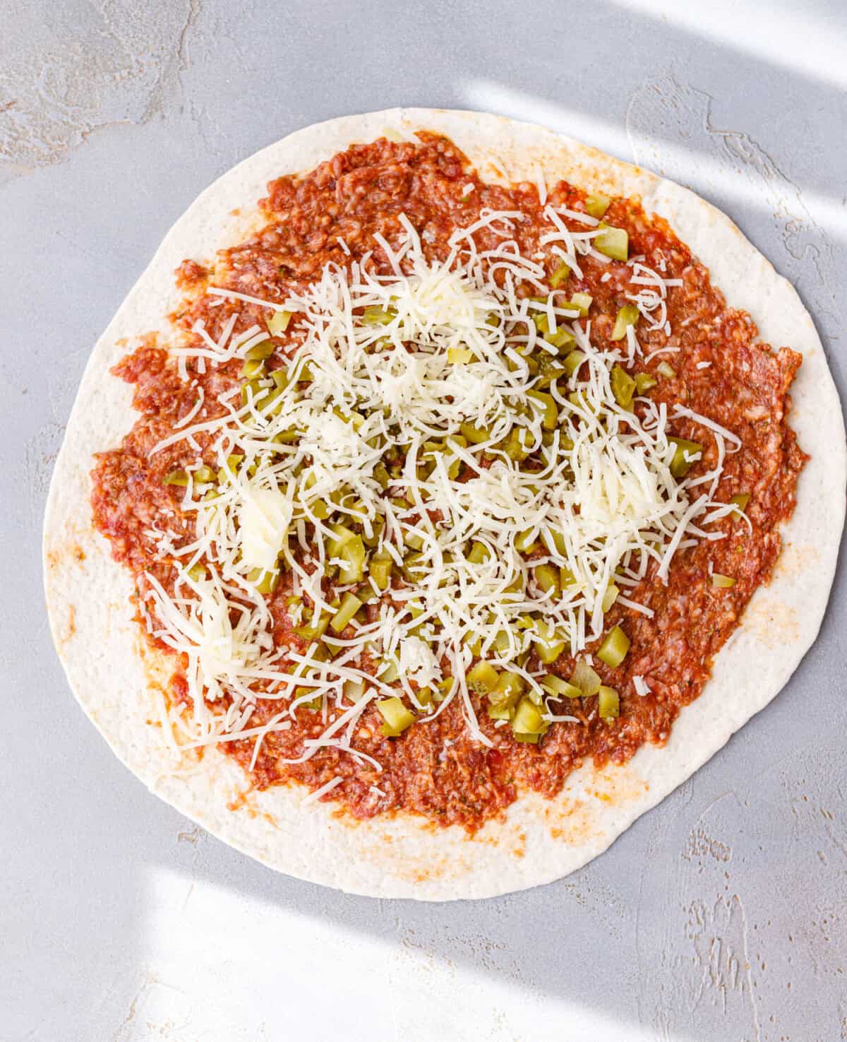 A flour tortilla spread with raw seasoned ground beef and topped with chopped pickles and grated cheese.