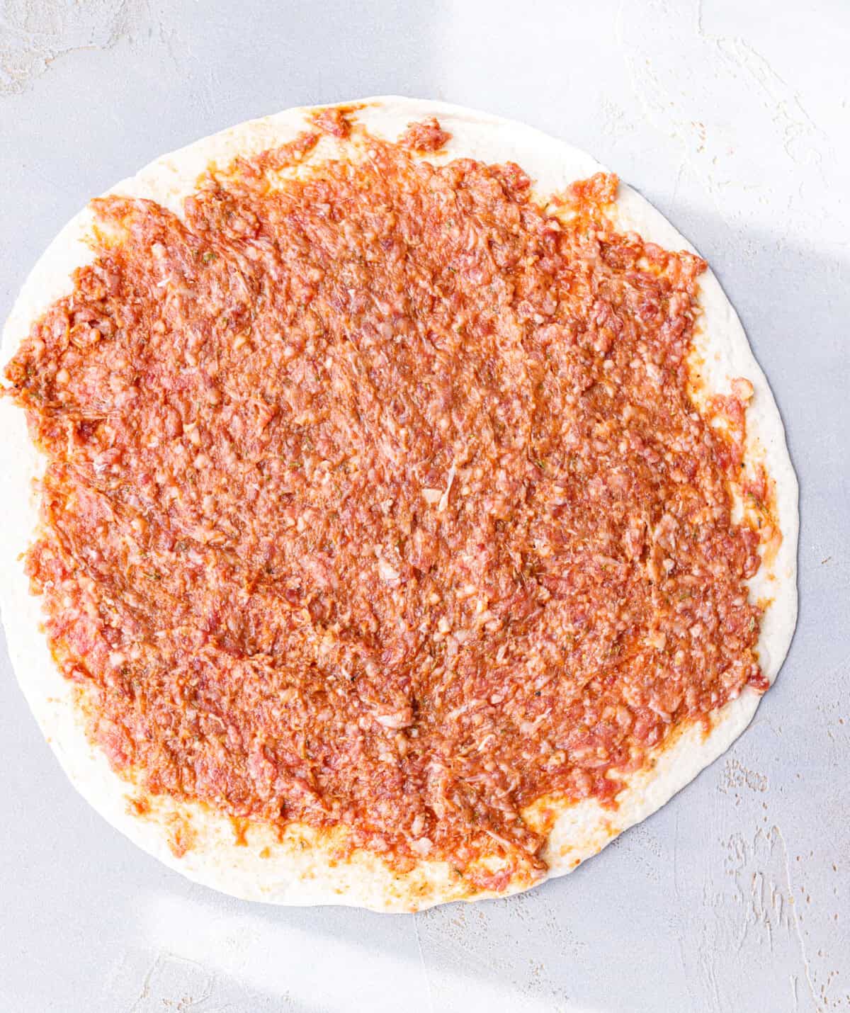 Raw seasoned ground beef spread in a thin even layer across a flour tortilla, right to the edges.
