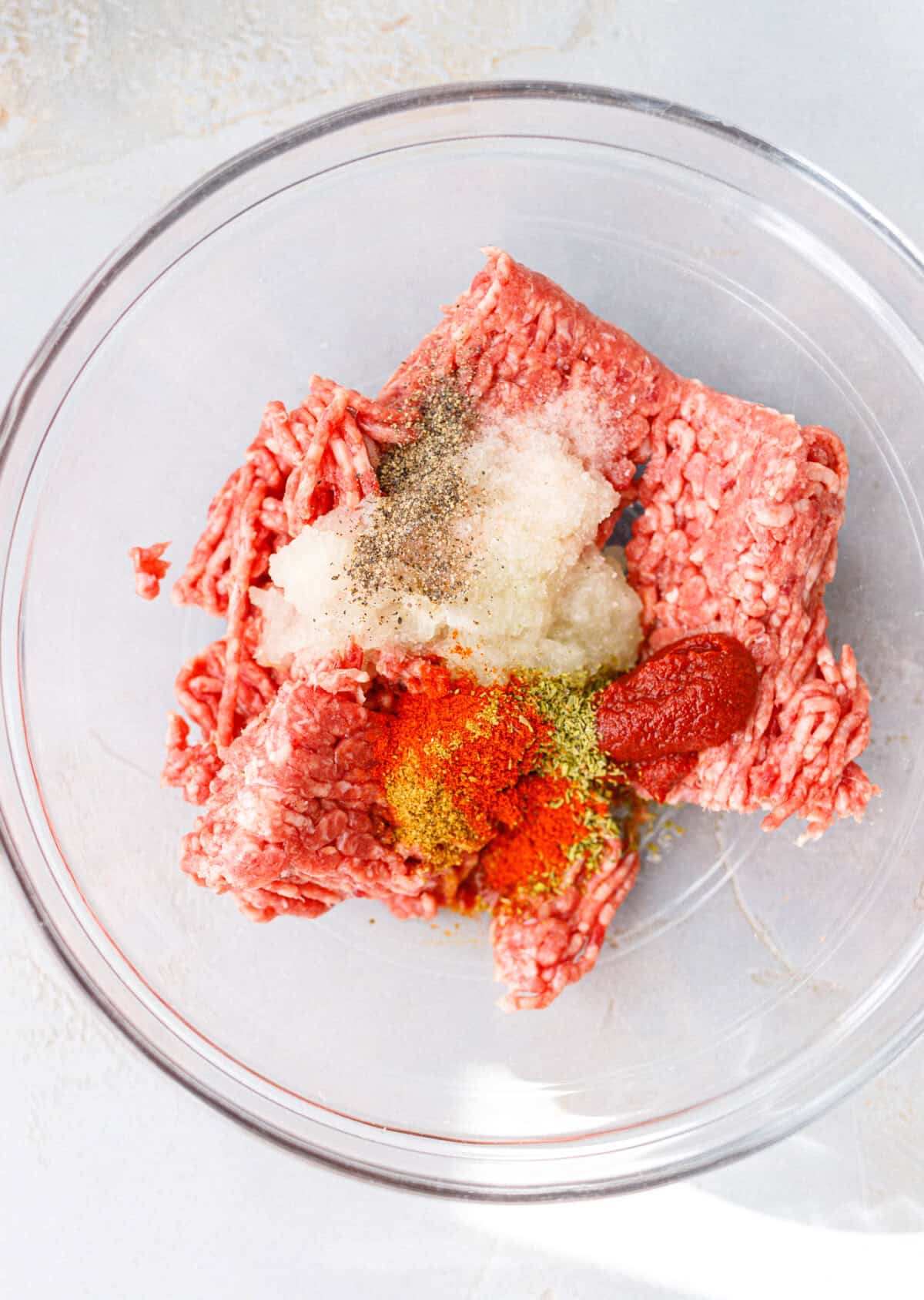 Raw ground beef in a glass bowl with grated onion, tomato paste, smoked paprika, cumin, chubritsa, salt and pepper before mixing.