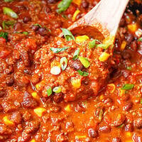 A wooden spoon lifting crockpot chili with ground beef, kidney and black beans, corn and green onion.