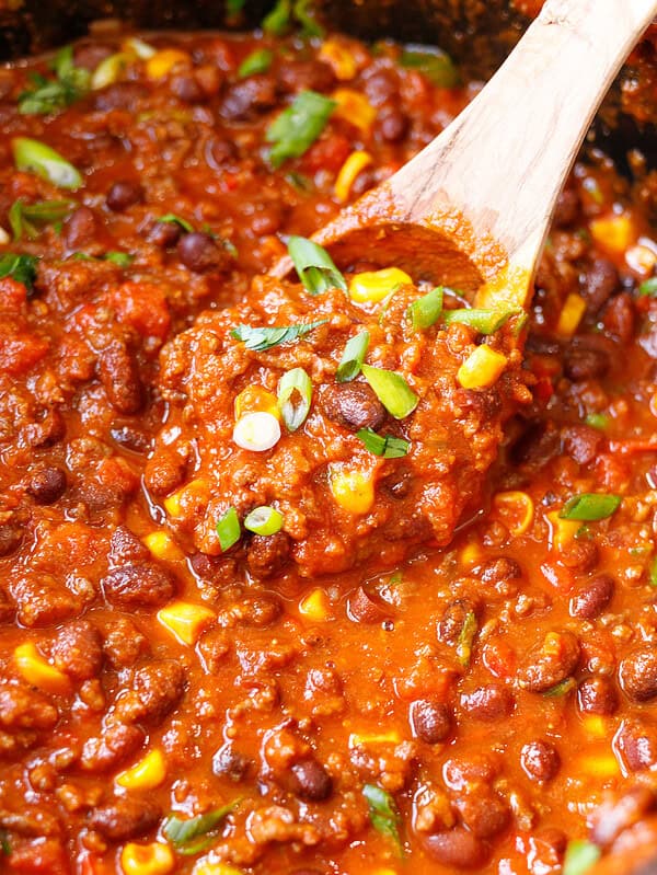 A wooden spoon lifting crockpot chili with ground beef, kidney and black beans, corn and green onion.