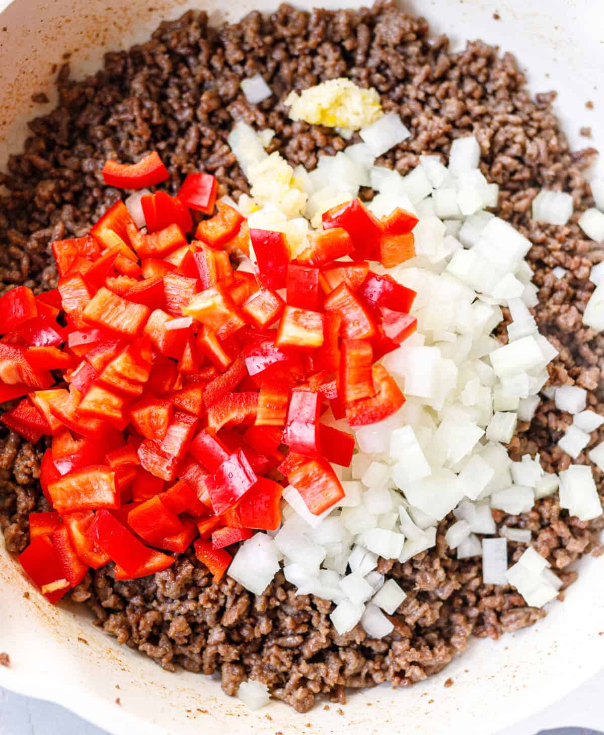 Diced red bell pepper, chopped onion and minced garlic added on top of browned ground beef in a skillet.