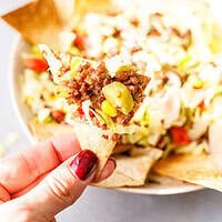A hand holding a homemade tortilla chip topped with ground beef taco salad, with the salad bowl behind it.