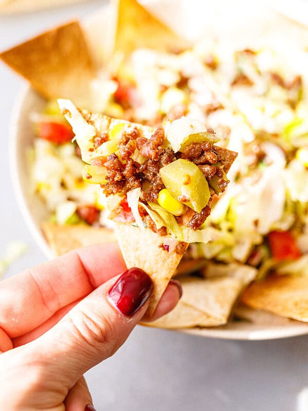 A hand holding a homemade tortilla chip topped with ground beef taco salad, with the salad bowl behind it.