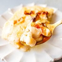 A gold spoon holding a bite of baked pumpkin custard with soft roasted pumpkin pieces, in front of a slice on a scalloped white plate.