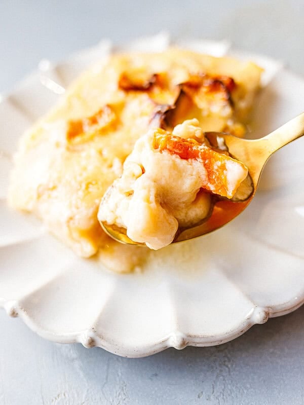 A gold spoon holding a bite of baked pumpkin custard with soft roasted pumpkin pieces, in front of a slice on a scalloped white plate.